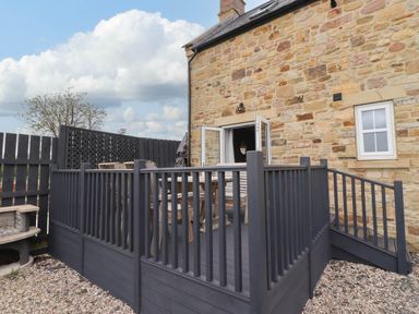 An outdoor patio with a dark wooden fence and wooden furniture next to a stone house at Oak Cottage in Lucker near Warenford