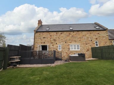 A stone house with a black wooden deck and patio furniture a hot tub and a grassy yard at Oak Cottage in Lucker near Warenford