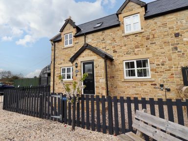 A stone house with a black front door and white windows behind a black wooden fence and gravel ground at Yew Cottage in Lucker near Warenford
