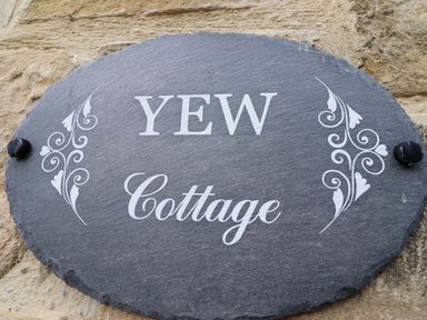 An oval slate house sign with white decorative patterns and the words Yew Cottage mounted on a stone wall at Yew Cottage in Lucker near Warenford