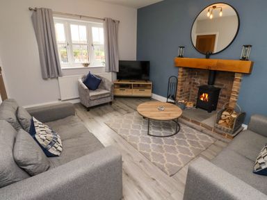 A living room with two gray sofas a gray armchair a round wooden coffee table a rug a TV on a wooden stand a fireplace with a round mirror above and wood logs at Yew Cottage in Lucker near Warenford
