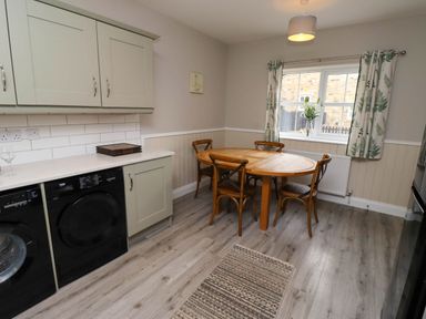 A kitchen with a round wooden dining table with four chairs a window with curtains and appliances under the countertop at Yew Cottage in Lucker near Warenford