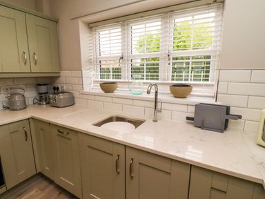 A kitchen with a sink a faucet two bowls a glass pitcher toasters a kettle a coffee maker and cabinets under a window at Yew Cottage in Lucker near Warenford