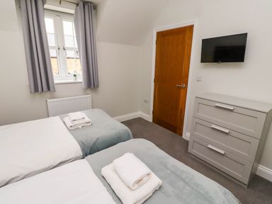 A bedroom with two single beds with folded towels on them a window with curtains a wooden door a gray chest of drawers and a wall-mounted television at Yew Cottage in Lucker near Warenford
