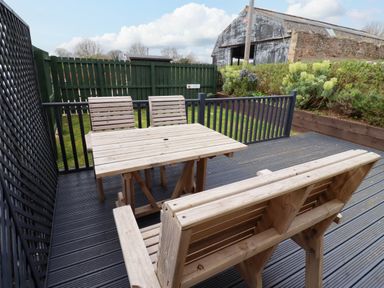 An outdoor wooden deck with a rectangular table two chairs and a bench with a green fence and a barn in the background at Yew Cottage in Lucker near Warenford