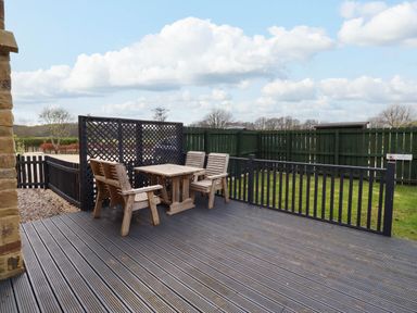 An outdoor deck with wooden table and chairs next to a green fenced lawn at Yew Cottage in Lucker near Warenford