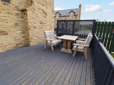 An outdoor patio area with wooden benches and a table on a gray deck at Yew Cottage in Lucker near Warenford