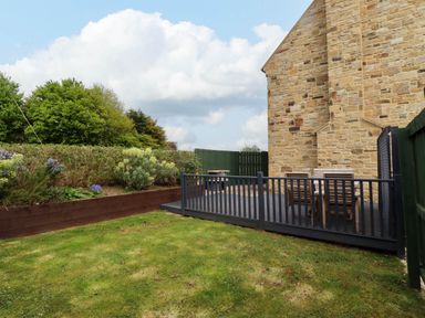 A garden with grass and shrubs next to a stone building and a deck with outdoor furniture at Yew Cottage in Lucker near Warenford
