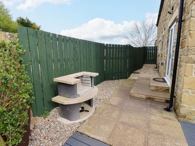 A stone patio with steps leading to a door and a concrete barbecue next to a green wooden fence at Yew Cottage in Lucker near Warenford