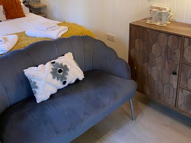 A room with a gray sofa with a patterned cushion a wooden cabinet with cups and glasses and a bed with towels and a yellow blanket at Red Squirrels Drey in Lucker near Belford