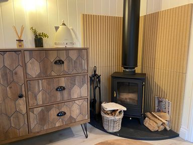 A wood stove with firewood and a wooden cabinet with a kettle and plant on top at Red Squirrels Drey in Lucker near Belford
