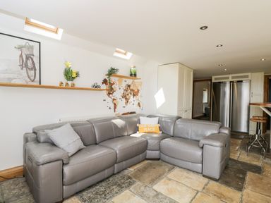 A living room with a gray leather sectional sofa under skylights and a wall shelf displaying decorations at 2 Dewars Farm Cottages in Middleton Stoney