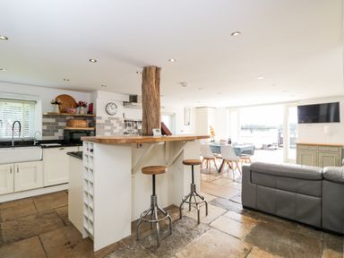 An open kitchen and dining area with a wooden breakfast bar and stools a dining table and a gray sofa at 2 Dewars Farm Cottages in Middleton Stoney