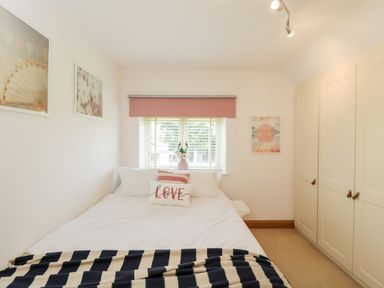 A bedroom with a bed with pillows and a blanket a window with blinds and a pink valance wardrobes and framed pictures on the walls at 2 Dewars Farm Cottages in Middleton Stoney