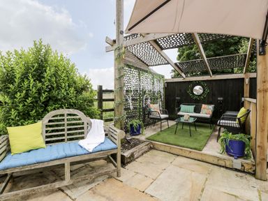 An outdoor seating area with wooden bench and cushioned metal chairs under a pergola at 2 Dewars Farm Cottages in Middleton Stoney