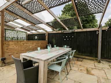 An outdoor patio dining area with a wooden table, several chairs, a pergola with patterned shade panels, and a black fence at 2 Dewars Farm Cottages Middleton Stoney