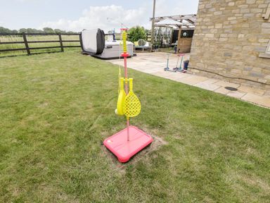 A backyard with a grass lawn and a badminton set with yellow rackets and a red base near a stone house with a paved patio and scooters at 2 Dewars Farm Cottages in Middleton Stoney