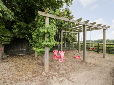An outdoor wooden play structure with swings and climbing rings near trees and a fence at 2 Dewars Farm Cottages in Middleton Stoney