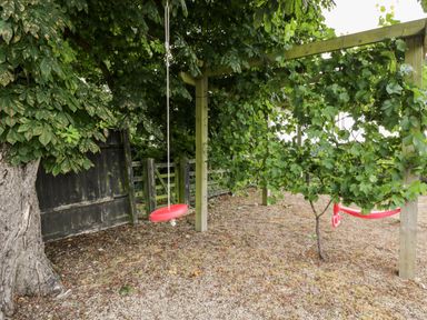An outdoor area with two red swings hanging from a wooden structure surrounded by trees and greenery at 2 Dewars Farm Cottages in Middleton Stoney
