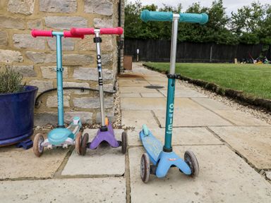 Three children's scooters standing on a stone patio next to a stone wall with a blue planter and grass in the background at 2 Dewars Farm Cottages Middleton Stoney