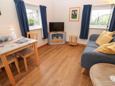 A living room with a blue sofa and yellow pillows a wooden dining table two windows with blue curtains and a television on a gray stand at Ramblers Rest in St Just