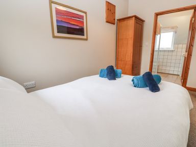 A bedroom with a bed covered in white bedding and folded towels on top next to a wooden wardrobe and an open door leading to a tiled bathroom at Ramblers Rest in St Just