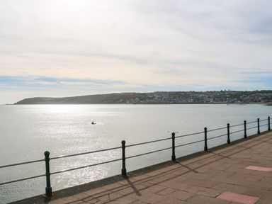 A waterfront promenade with metal railings overlooking a calm sea and a distant coastline with houses at Ramblers Rest in St Just
