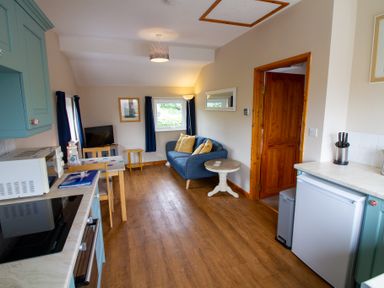 A kitchen area with blue cabinets a microwave and stove a small dining table and chairs a blue sofa with pillows a white round side table and wooden door at Ramblers Rest in St Just