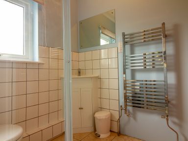 A bathroom with a shower enclosure a small sink cabinet a white trash bin a chrome towel radiator and a window at Ramblers Rest in St Just