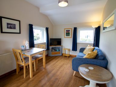 A living room with a blue sofa yellow cushions wooden dining table chairs and a TV at Ramblers Rest in St Just
