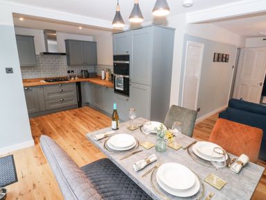 A kitchen with gray cabinets and wooden counters next to a dining table set with plates glasses and cutlery at Rose Cottage in Bishopsteignton