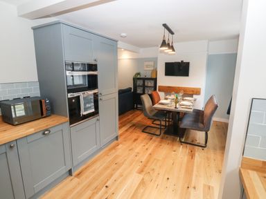 A kitchen and dining area with grey cabinets wooden floors dining table set with plates and glasses and a wall mounted television at Rose Cottage in Bishopsteignton