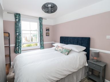 A bedroom with a white bed and dark blue headboard next to a window with patterned curtains at Rose Cottage in Bishopsteignton
