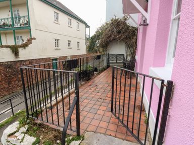 A tiled patio with black railings and an open gate next to a pink building with outdoor seating and a vine-covered area at Rose Cottage in Bishopsteignton