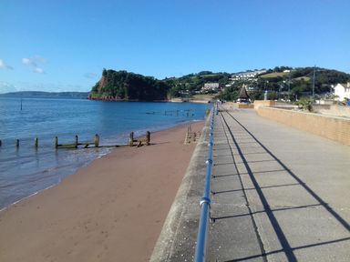 A beach with a promenade and a railing along the side with hills and buildings in the background at Rose Cottage in Bishopsteignton