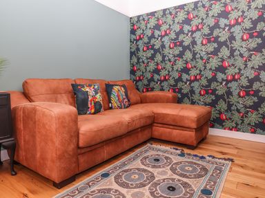 A brown leather sofa with two colorful animal print cushions next to a side table floral wallpaper on one wall wooden floor with a patterned rug at Rose Cottage in Bishopsteignton