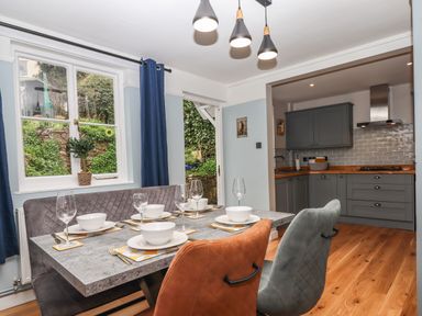 A dining area with a stone table set with dishes and glasses next to a kitchen with grey cabinets and wooden floor at Rose Cottage in Bishopsteignton