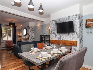 A dining table set with bowls and wine glasses in an open living and dining room with patterned wallpaper and a wall-mounted TV at Rose Cottage in Bishopsteignton