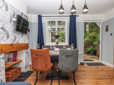 A dining area with mixed color chairs a table set with glasses a television on the wall and a door leading outside at Rose Cottage in Bishopsteignton