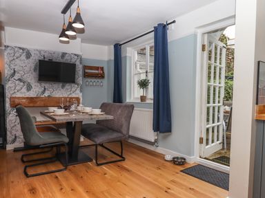 A dining area with a table set for six, chairs, a wall-mounted television, blue curtains, and a door leading outside at Rose Cottage in Bishopsteignton