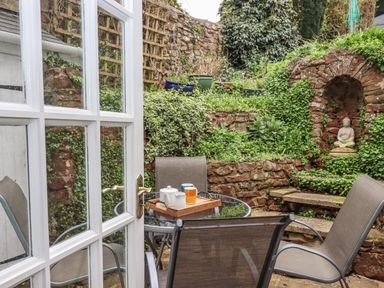 A garden patio with a glass table and four chairs a tray with a teapot and cups on the table stone steps and a Buddha statue in a niche surrounded by greenery at Rose Cottage in Bishopsteignton