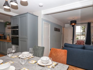 A dining table set with white bowls and wine glasses next to a kitchen with grey cabinets and an adjoining living area with blue sofa and window at Rose Cottage in Bishopsteignton