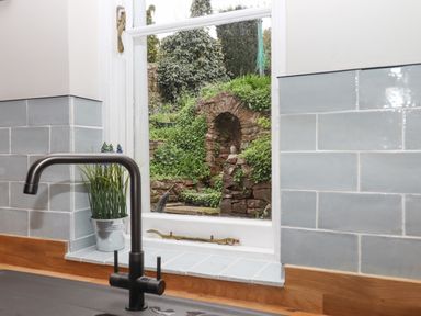 A kitchen sink with a black faucet and potted plant by a window showing a garden with a stone arch and greenery at Rose Cottage in Bishopsteignton
