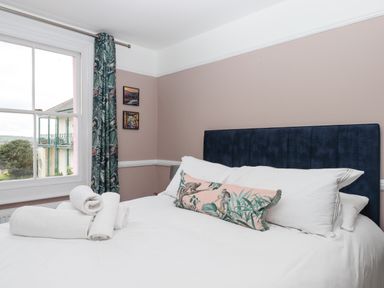 A bedroom with a bed with white linens and a decorative pillow near a window with patterned curtains at Rose Cottage in Bishopsteignton