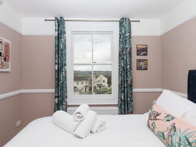 A bedroom with a bed with white linens and rolled towels by a window with patterned curtains at Rose Cottage in Bishopsteignton