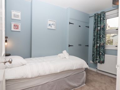 A bedroom with a single bed with white bedding blue walls framed pictures a window with patterned curtains and built in cupboards at Rose Cottage in Bishopsteignton