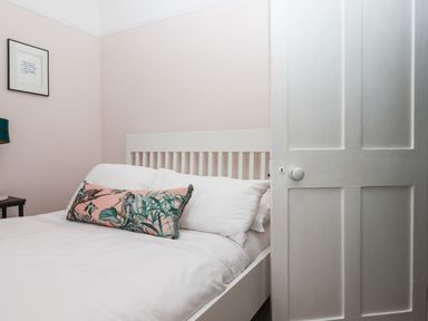 A bedroom with a white wooden bed a pink decorative pillow a nightstand and a partially opened white door at Rose Cottage in Bishopsteignton