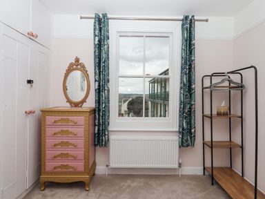 A bedroom with a window curtains a pink and gold chest of drawers and a black metal clothing rack at Rose Cottage in Bishopsteignton