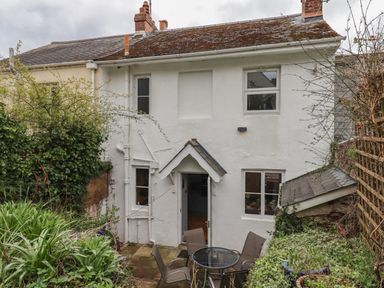 The rear exterior of a white two-story house with a small patio and garden area with plants and outdoor furniture at Rose Cottage in Bishopsteignton