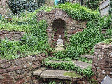 A stone garden with green plants and a Buddha statue in a stone alcove at Rose Cottage in Bishopsteignton